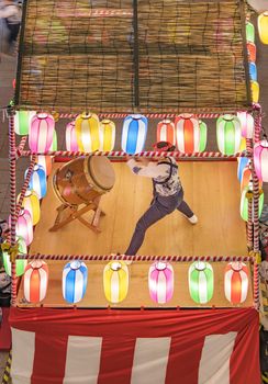 View of the square in front of the Nippori train station decorated for the Obon festival with a yagura tower illuminated with paper lanterns where a girl in traditional costume is playing taiko drum in the summer night of Arakawa district in Tokyo.