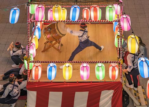 View of the square in front of the Nippori train station decorated for the Obon festival with a yagura tower illuminated with paper lanterns where a girl in traditional costume is playing taiko drum in the summer night of Arakawa district in Tokyo.