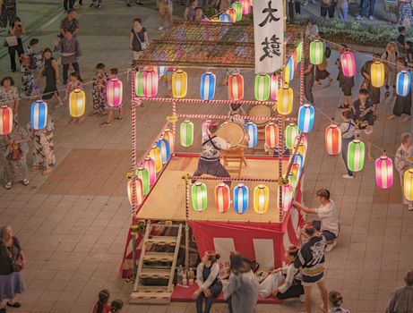 View of the square in front of the Nippori train station decorated for the Obon festival with a yagura tower illuminated with paper lanterns where a girl in traditional costume is playing taiko drum in the summer night of Arakawa district in Tokyo.