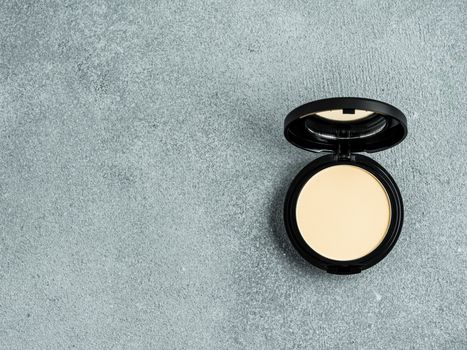 Compact powder on gray cement background. Female pressed powder in opened black plastic case with mirror, copy space for text or design. Top view or flat lay