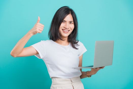 Asian beautiful young woman smile white teeth standing to holding laptop computer and showing thumb up while shoot photo in studio on blue background with copy space for text