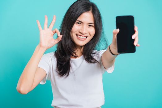 Portrait Asian beautiful happy young woman standing smile, holding mobile phone and showing ok gesture, shoot photo in studio focus face on blue background, with copy space