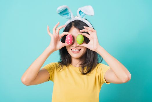 Portrait Asian beautiful happy young woman smile white teeth wear yellow t-shirt standing with bunny ears and holding Easter eggs near the eye, on blue background with copy space