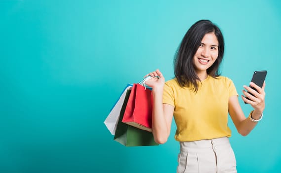 Portrait happy Asian beautiful young woman smile white teeth standing wear yellow t-shirt, She holding shopping bags and using a mobile phone, studio shot on blue background with copy space for text