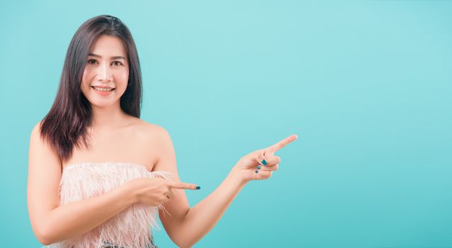 Asian happy portrait beautiful young woman standing her pointing finger outside and looking to camera isolated on blue background with copy space for text