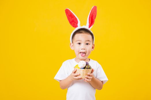 Portrait happy Asian cute little children boy smile standing so happy wearing white T-shirt and bunny ears in Easter festival day holding easter eggs, studio shot on yellow background with copy space