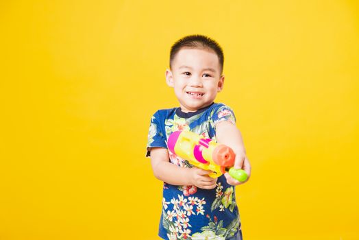 Portrait happy Asian cute little children boy smile standing so happy wearing flower shirt in Songkran festival day holding water gun, studio shot on yellow background with copy space