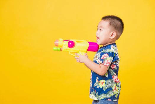 Portrait happy Asian cute little children boy smile standing so happy wearing flower shirt in Songkran festival day holding water gun, studio shot on yellow background with copy space