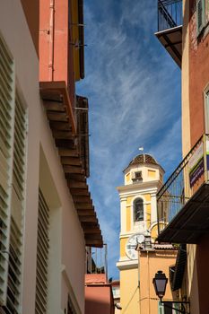 Yellow plaster clock tower rising past narrow street under blue sky in Villefranche, France