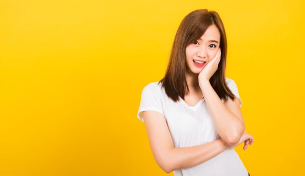 Asian happy portrait beautiful cute young woman teen standing wear white t-shirt posting rest chin on hand looking to camera isolated, studio shot on yellow background with copy space for text