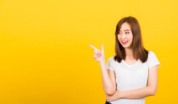 Asian happy portrait beautiful cute young woman teen standing wear t-shirt pointing finger away side looking to side isolated, studio shot on yellow background with copy space