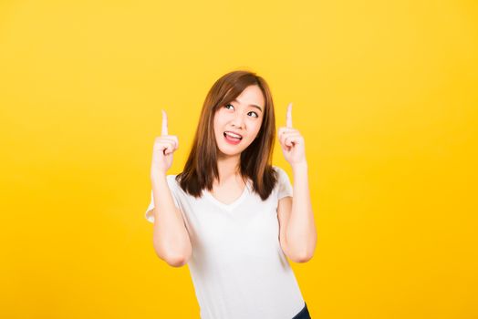 Asian happy portrait beautiful cute young woman teen standing wear t-shirt makes gesture two fingers point upwards above looking to camera isolated, studio shot on yellow background with copy space