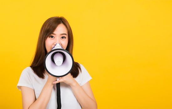 Asian happy portrait beautiful cute young woman teen standing making announcement message shouting screaming in megaphone looking to camera isolated, studio shot on yellow background with copy space