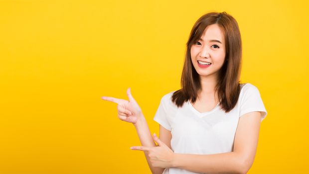 Asian happy portrait beautiful cute young woman teen standing wear t-shirt pointing finger away side looking to camera isolated, studio shot on yellow background with copy space