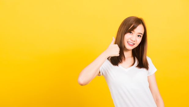 Asian happy portrait beautiful cute young woman teen standing wear t-shirt showing gesturing finger thumb up looking to camera isolated, studio shot on yellow background with copy space