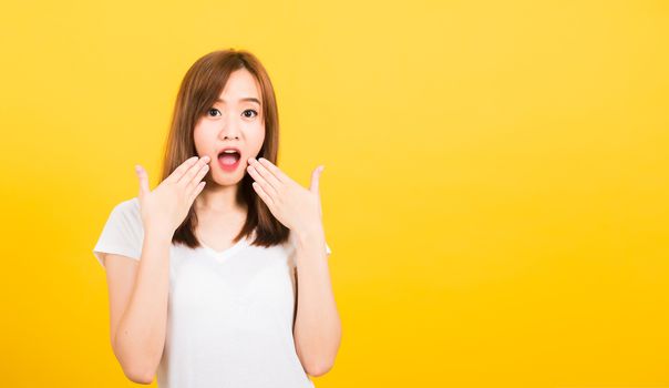 Asian happy portrait beautiful cute young woman teen standing amazed, shocked afraid wide open mouth eyes gesturing palms looking to camera isolated, studio shot on yellow background with copy space