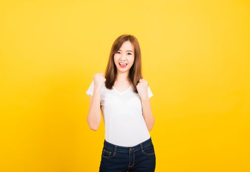 Asian happy portrait beautiful cute young woman teen standing wear t-shirt makes raised fists up celebrating her success looking to camera isolated, studio shot on yellow background with copy space