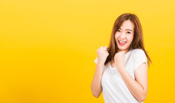 Asian happy portrait beautiful cute young woman teen standing wear t-shirt makes raised fists up celebrating her success looking to camera isolated, studio shot on yellow background with copy space