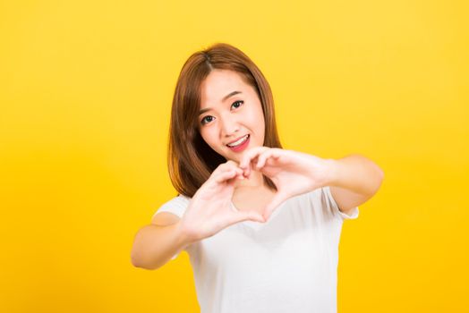 Asian happy portrait beautiful cute young woman teen smile standing make finger heart figure symbol shape sign with two hands looking to camera isolated, studio shot yellow background with copy space