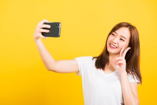 Asian happy portrait beautiful cute young woman teen smiling standing wear t-shirt making selfie photo, video call on smartphone looking camera isolated, studio shot yellow background with copy space