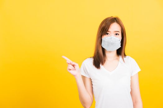 Portrait Asian beautiful happy young woman wearing face mask protects filter dust pm2.5 anti-pollution, anti-smog, and air pollution her pointing out to side on yellow background, with copy space