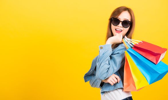 Asian happy portrait beautiful cute young woman teen smiling standing with sunglasses excited holding shopping bags multi color looking camera isolated, studio shot yellow background with copy space