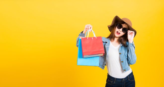 Asian happy portrait beautiful cute young woman teen smiling standing with sunglasses excited holding shopping bags multi color looking camera isolated, studio shot yellow background with copy space