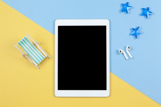 Tablet, wireless headphones, deck chair on yellow and blue background, starfishes, copy space, flat lay. Working space of freelancer. Freelance, planning of vacation, travel concept. Horizontal.