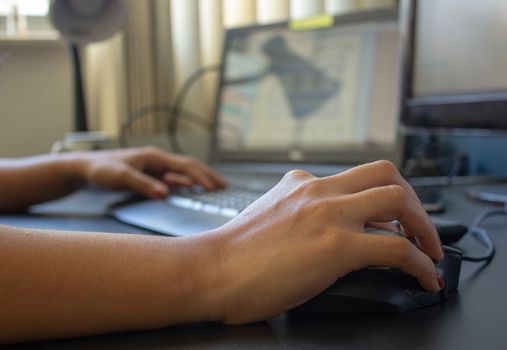 Closeup on the hand of a woman using her computer, with a keyboard, laptop and monitor on the background