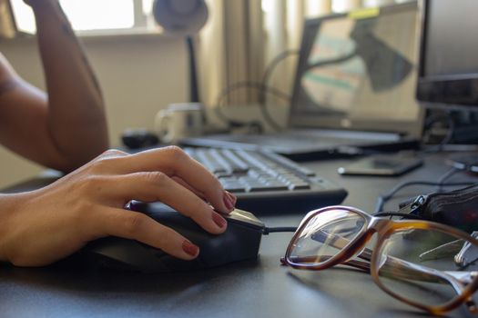 Closeup on the hand of a woman using her computer and glasses, with a keyboard, laptop and monitor on the background