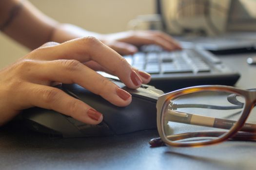 Closeup on the hand of a woman using her computer and glasses, with a keyboard, laptop and monitor on the background