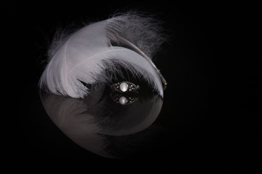 An extreme close-up / macro photograph of a detail of a soft white feather, black background. With wedding ring