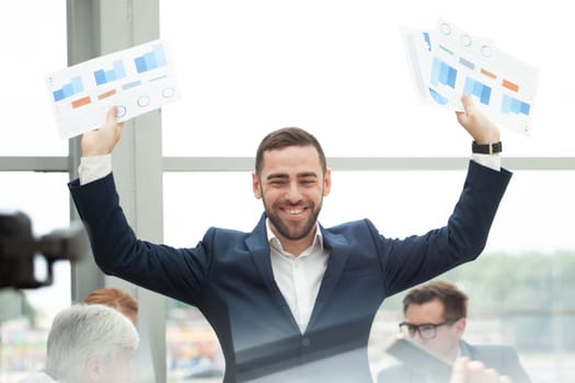 Young joyful businessman standing with arms raised holding data statistics diagrams and smiling , team of his colleagues sitting at the table in the background