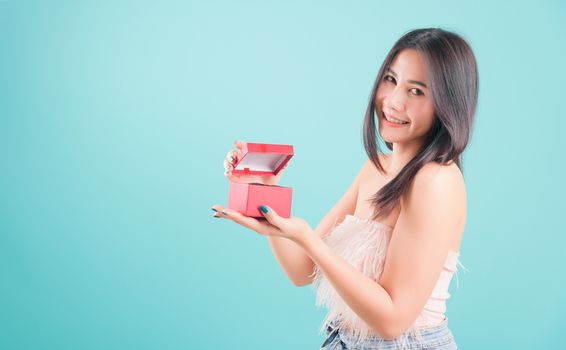Asian happy portrait beautiful young woman standing smile opening a red gift box, surprise birthday or Christmas day on blue background with copy space for text, healthy lifestyle concept