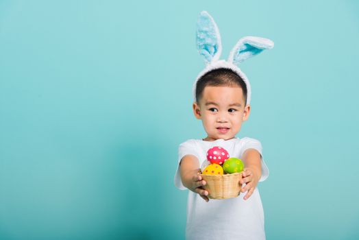 Asian cute little child boy smile beaming wearing bunny ears and a white T-shirt, standing to hold a basket with full Easter eggs on blue background with copy space