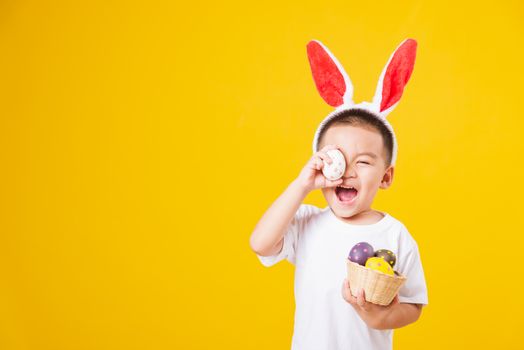 Portrait happy Asian cute little children boy smile standing so happy wearing white T-shirt and bunny ears in Easter festival day holding easter eggs, studio shot on yellow background with copy space