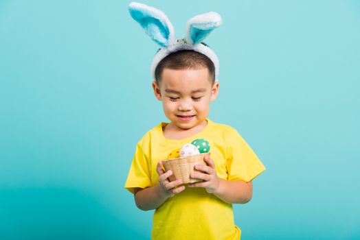 Asian cute little child boy smile beaming wearing bunny ears and a yellow T-shirt, standing to hold a basket with full Easter eggs on blue background with copy space