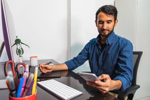 Young bearded businessman in denim shirt is sitting in office at table and is using computer looking at his smartphone. Man working