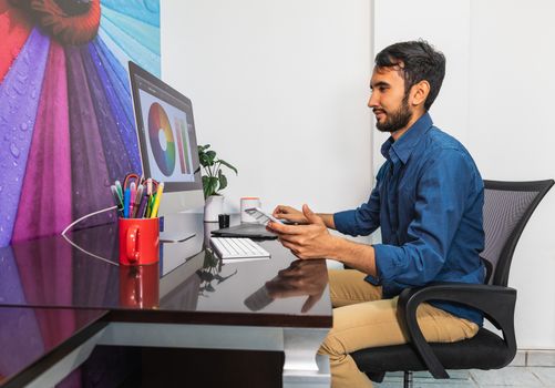 Side view. Young bearded businessman in denim shirt is sitting in office at table and is using computer. On table is smartphone and stationery. Man working