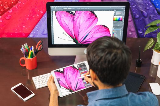 Back view. Young bearded businessman in denim shirt is sitting in office at table and is using a tablet. On table is smartphone and stationery. Man working