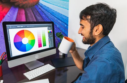 Side view. Smiling young bearded businessman in denim shirt is sitting in office at table. Drinking coffee. On computer color swatch