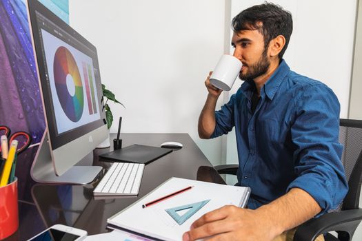 Side view. Smiling young bearded businessman in denim shirt is sitting in office at table. Drinking coffee. On computer color swatch