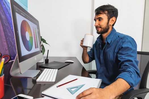 Side view. Smiling young bearded businessman in denim shirt is sitting in office at table. Drinking coffee. On computer color swatch