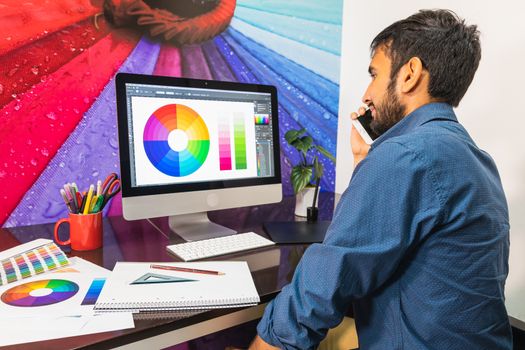 Side view. Young bearded businessman in denim shirt is sitting in office at table. Using his smartphone while looking at color swatch.