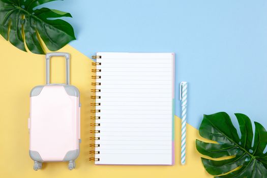 Notebook, pen, suitcase, monstera leaves on yellow and blue background, copy space, flat lay. For lifestyle blog, social media. Freelance, planning of vacation, travel, sales concept. Horizontal.