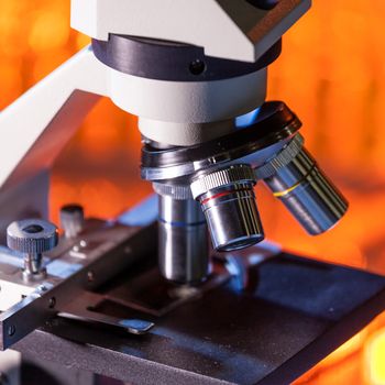 Close up of microscope lenses focused on a specimen in warm orange light light. Laboratory flask in the background.