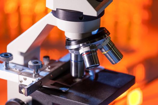 Close up of microscope lenses focused on a specimen in warm orange light light. Laboratory flask in the background.