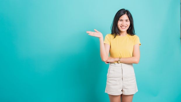 Portrait Asian beautiful young woman standing wear yellow t-shirt, She showing hand to presenting product and looking at the camera, shoot photo in a studio on a blue background, There was copy space