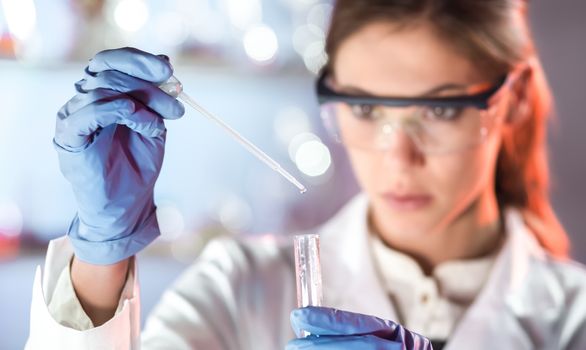 Life scientists researching in laboratory. Focused female life science professional pipetting solution into the glass cuvette. Lens focus on researcher's eyes. Healthcare and biotechnology concept.