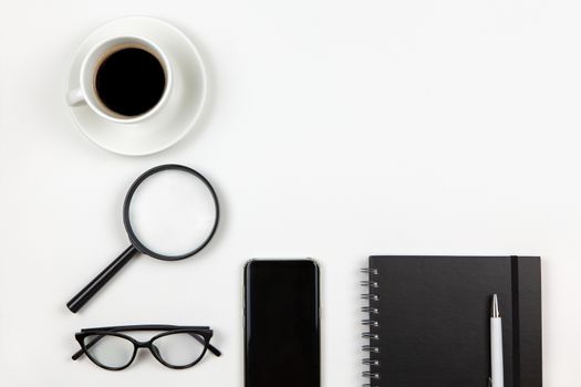 Modern unisex working space, top view. Notepad, pen, coffee, smartphone, magnifier, glasses on white background, copy space, flat lay. Desktop of freelancer. Work from home concept. Monochrome image.